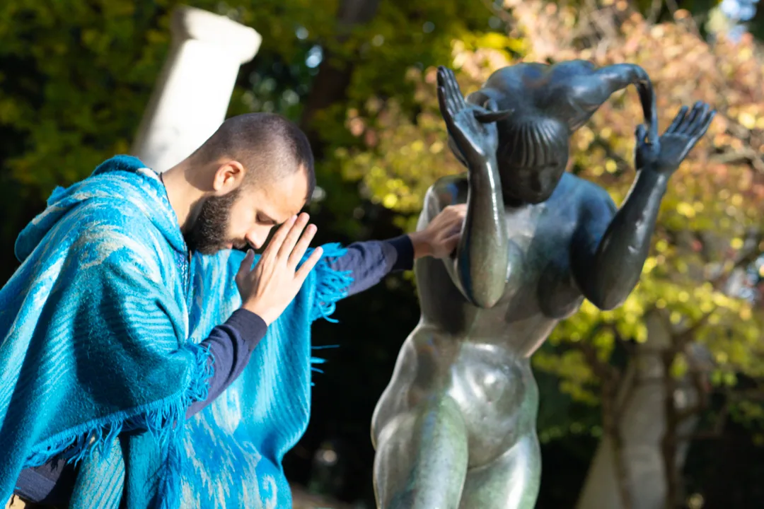 SANTOS in a blue cloak, with hands in prayer beside a sculpture