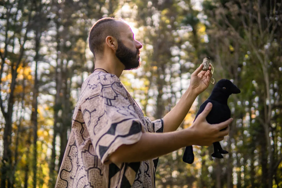 SANTOS holding burning sage and a crow figure in a woodland setting