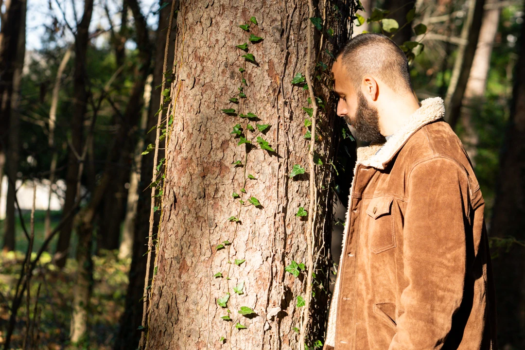 SANTOS resting his forehead against a tree in a sunlit woodland