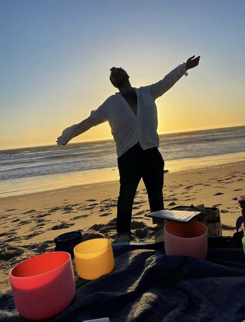 SANTOS standing beside singing bowls on the beach at sunset