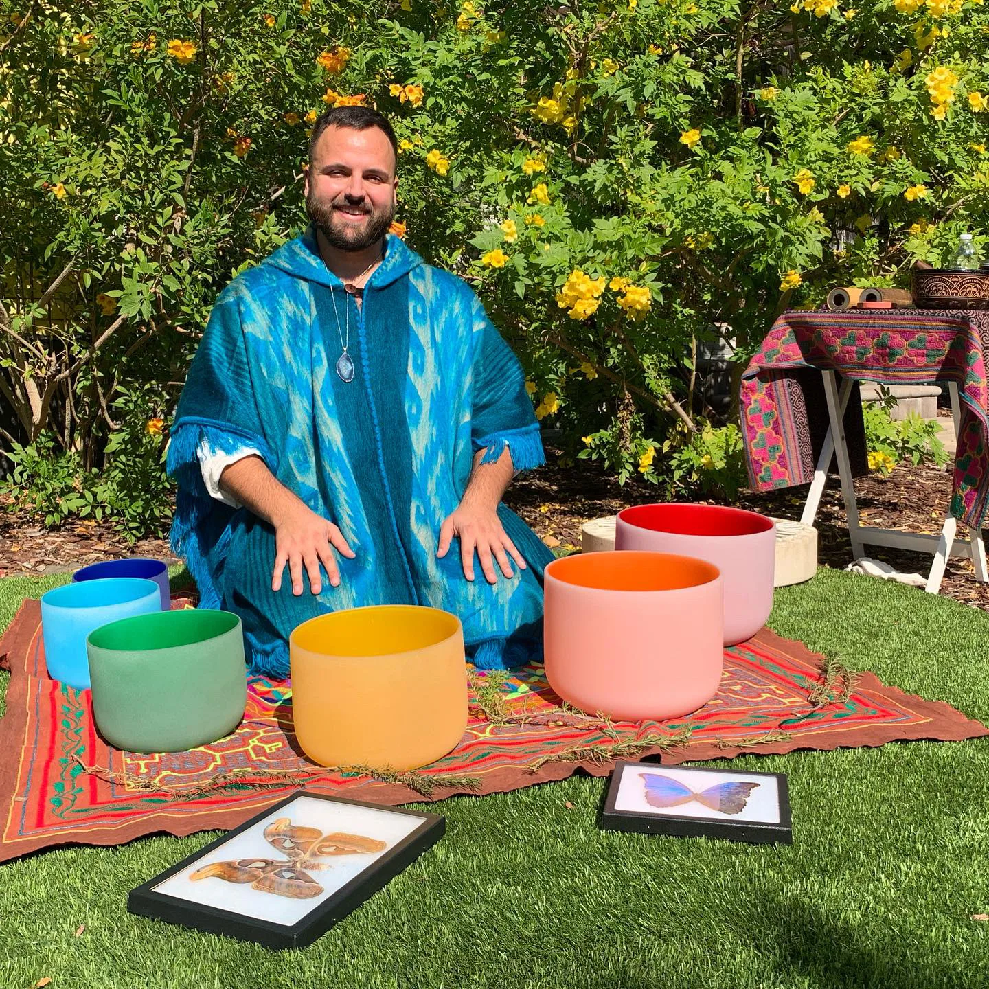 SANTOS in a blue cloak, seated behind colorful crystal singing bowls in a garden